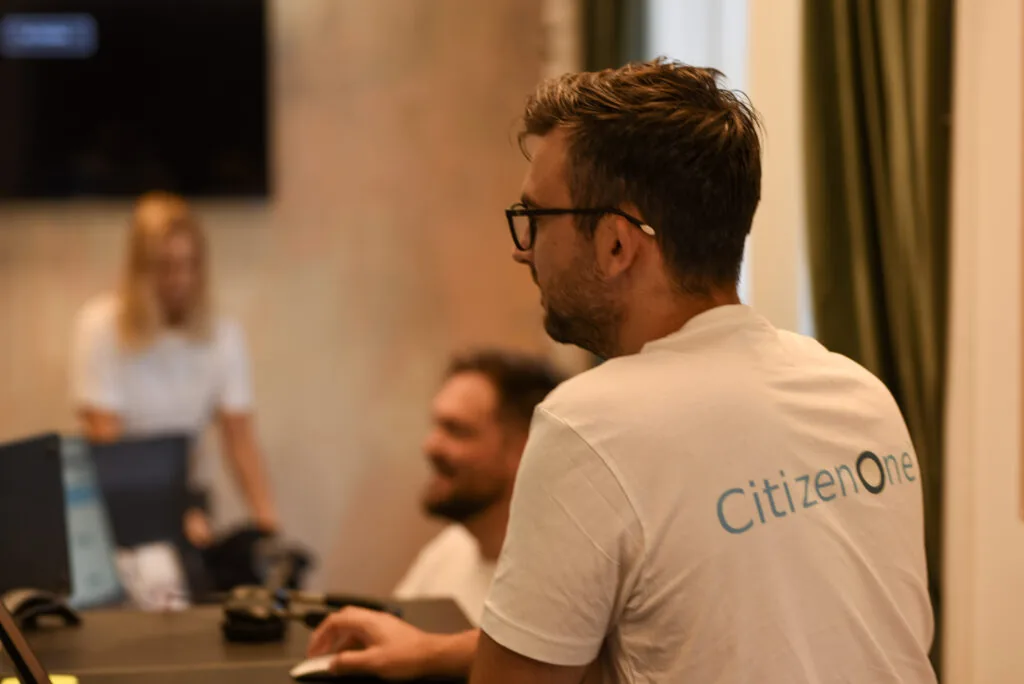 To medarbejdere hos CitizenOne ved deres skriveborde, den forreste i en CitizenOne-t-shirt.